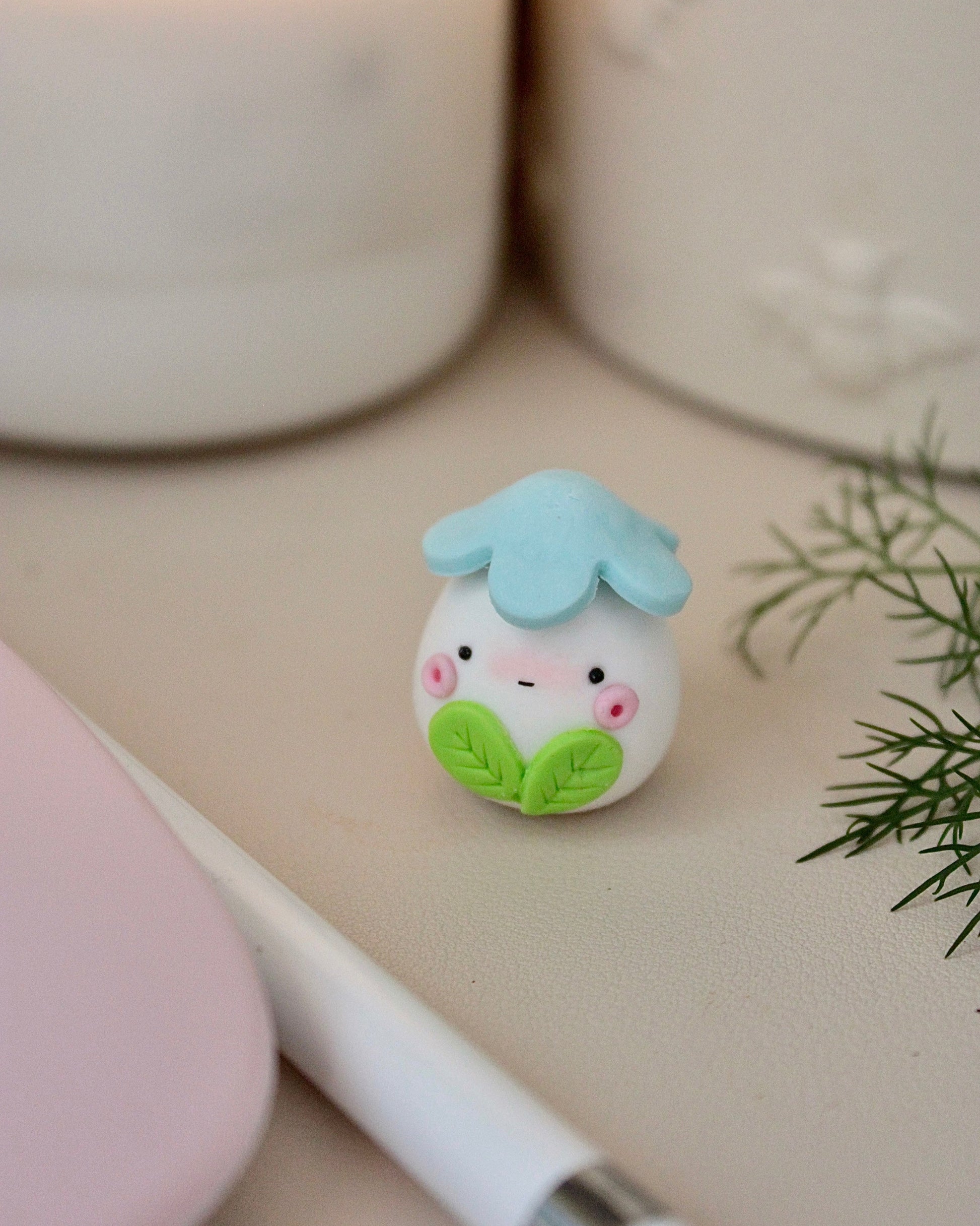 Small ceramic figurine with a blue flower on top, sitting on a surface with a pen and plant in the background.