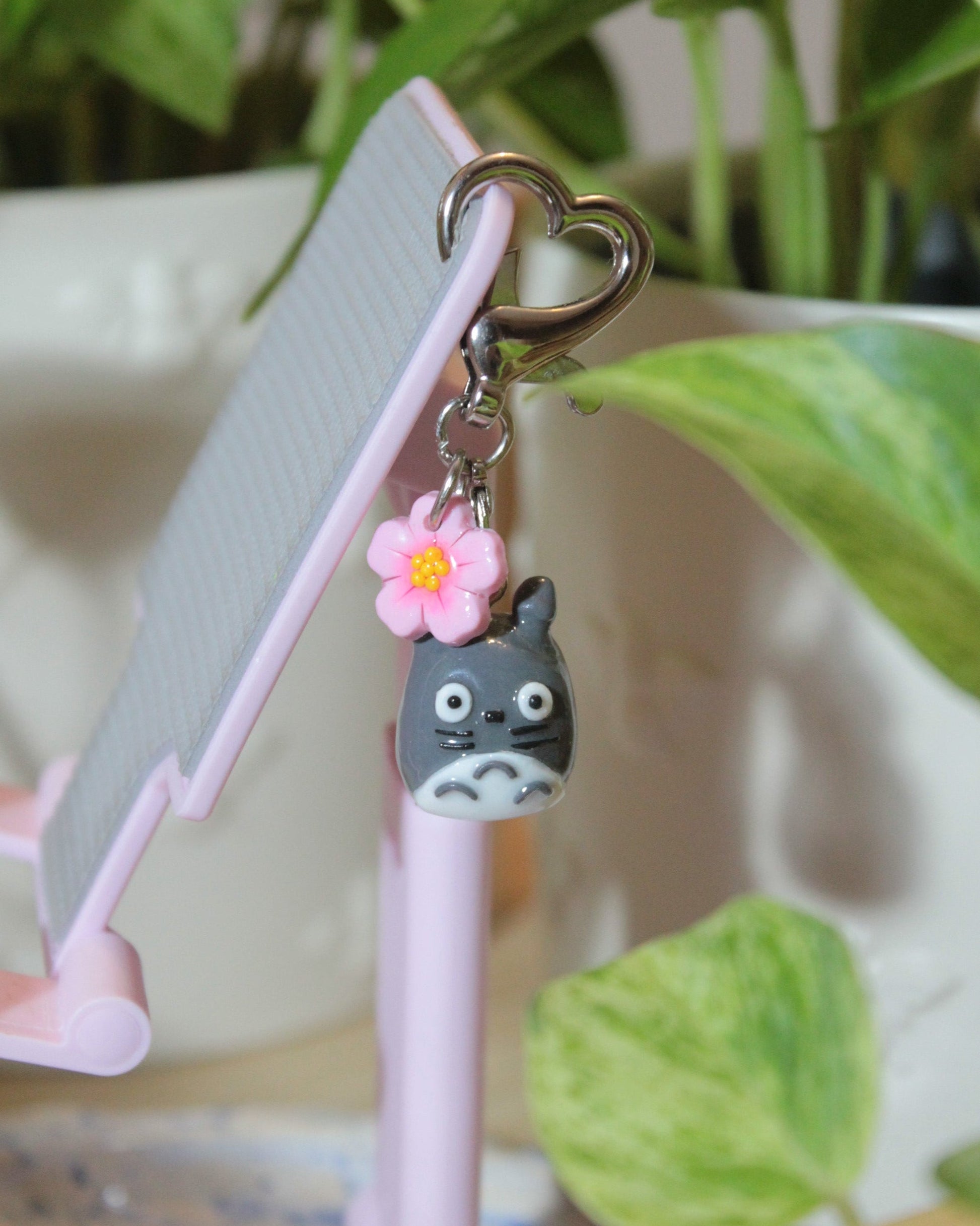 Pink phone stand with Totoro keychain and flower charm against a green leafy background